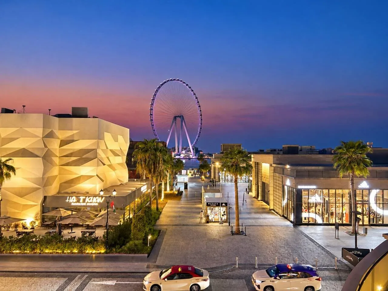 An outdoor shopping plaza at dusk, with a large illuminated Ferris wheel in the distance.