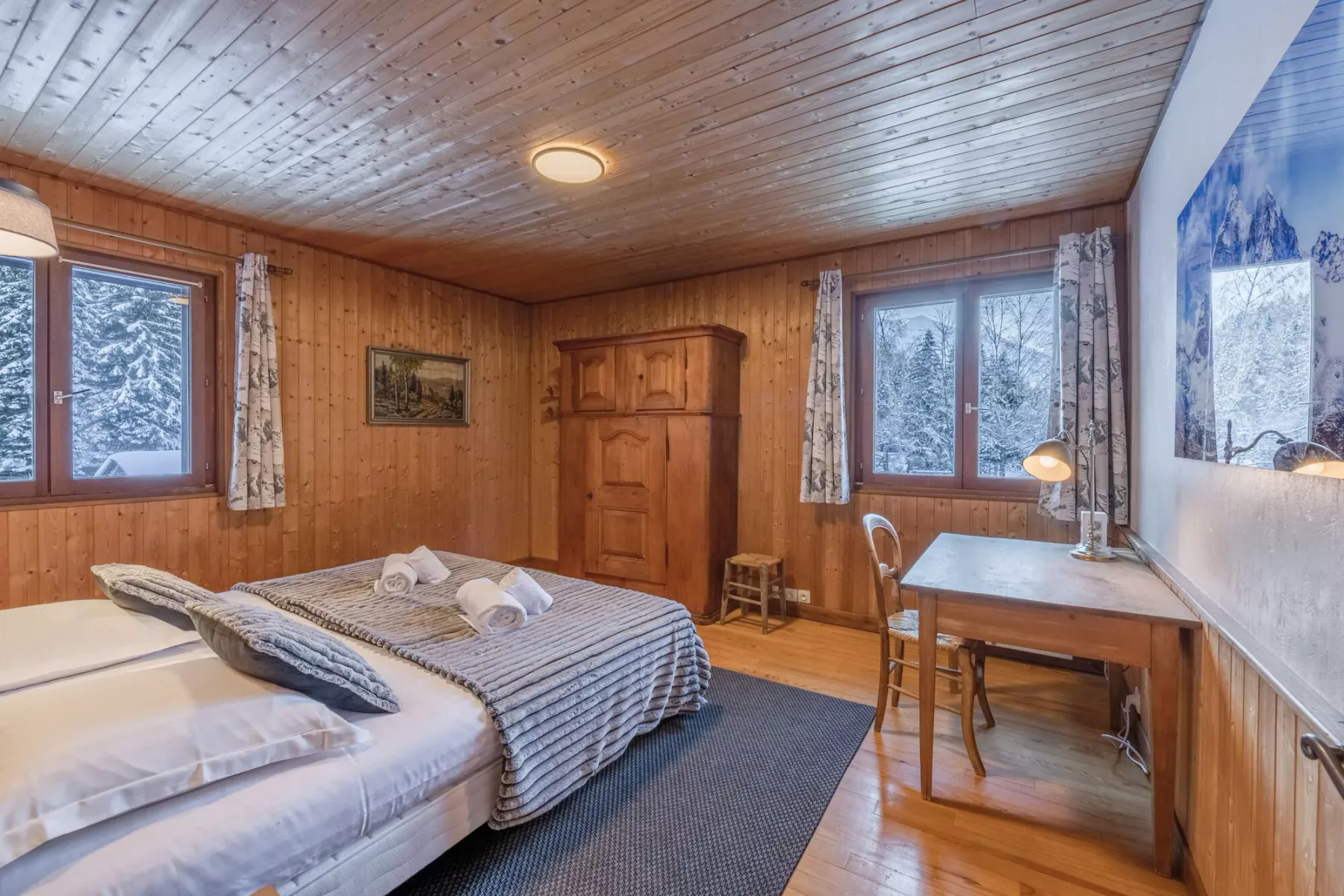 Un dormitorio revestido de madera con dos camas y un escritorio junto a las ventanas.
