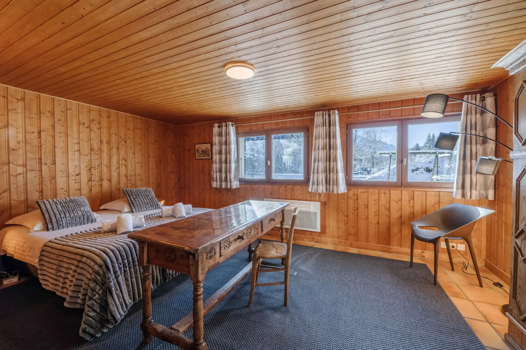 Habitación revestida de madera con una cama, una mesa de madera y ventanas con vistas a un paisaje nevado.