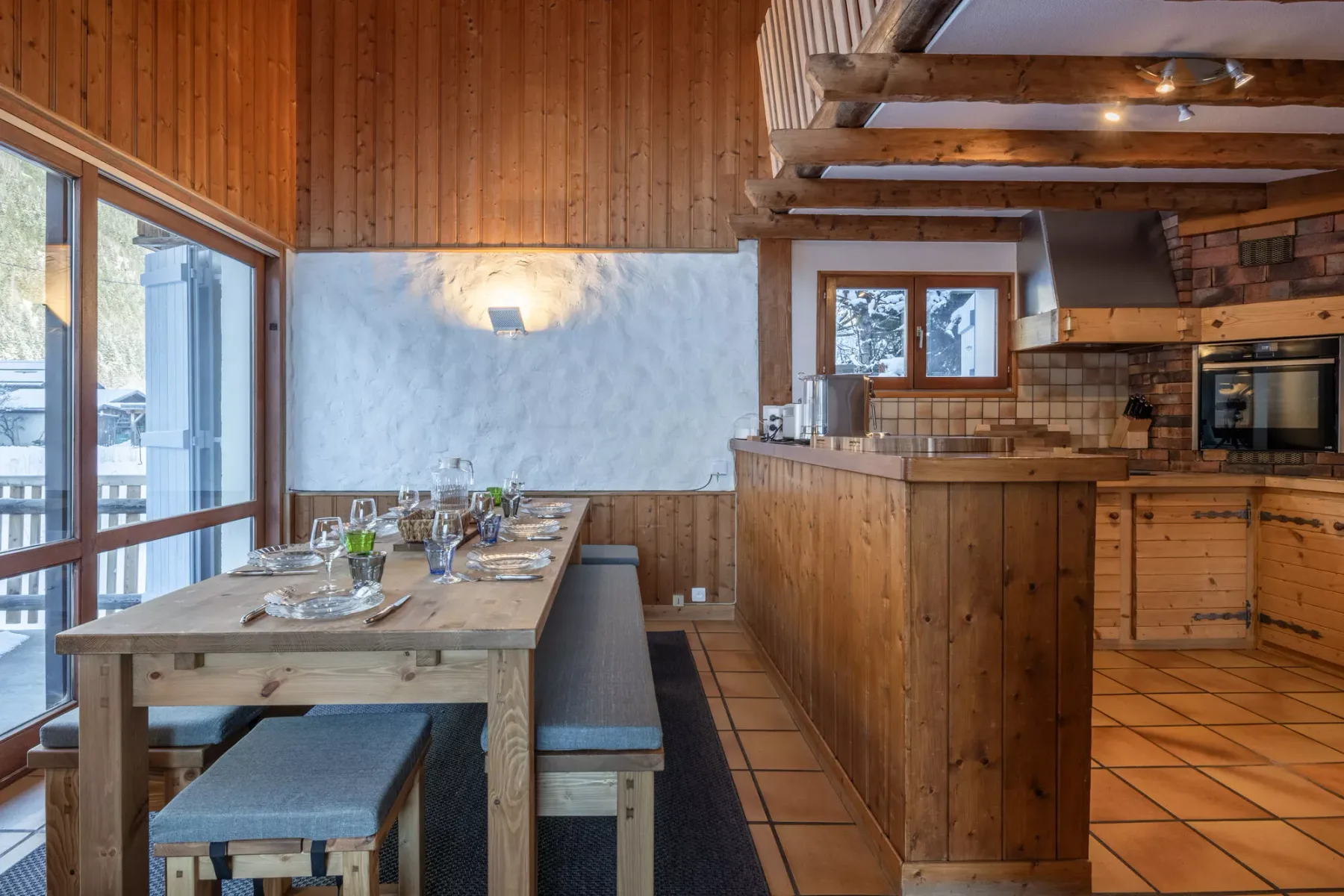 Comedor y cocina con una mesa de madera junto a grandes ventanas.