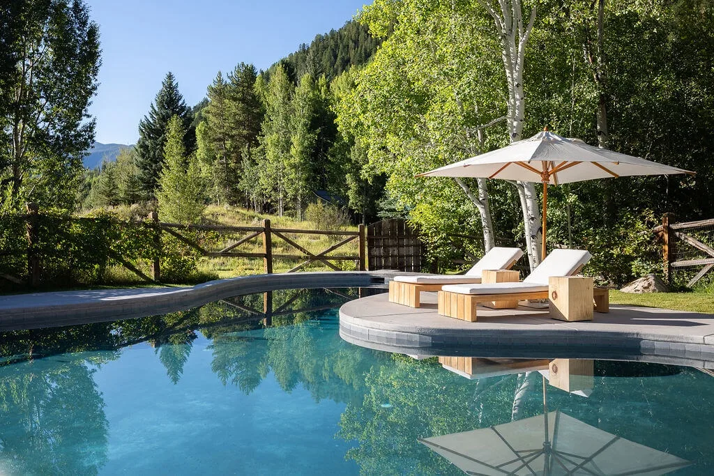 An outdoor pool beside a wooded landscape, with lounge seating under an umbrella.