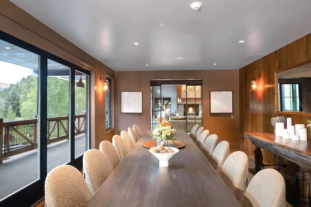 A dining room with a long table and chairs beside large windows.