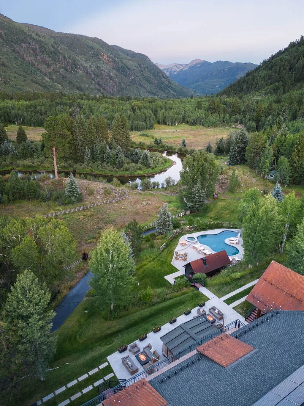 Aerial view of a mountain valley with a winding stream, forest, and swimming pool.