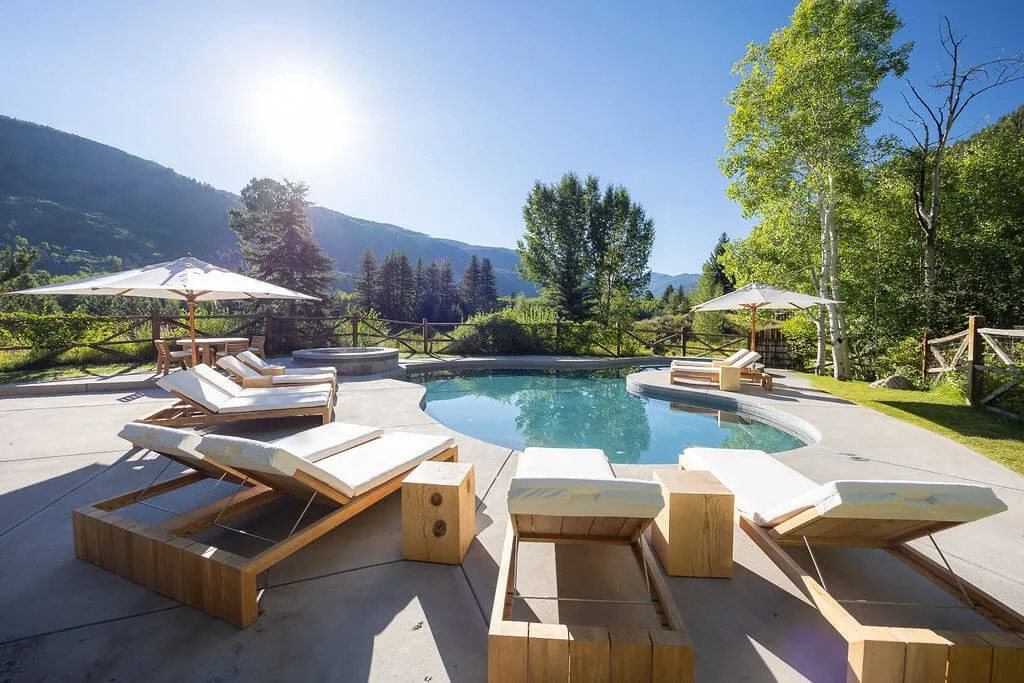 An outdoor pool sits beside a paved patio with lounge seating and umbrellas.