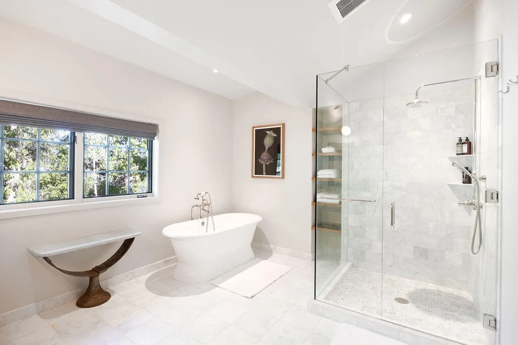 A bright bathroom features a freestanding bathtub beside a glass shower and window.