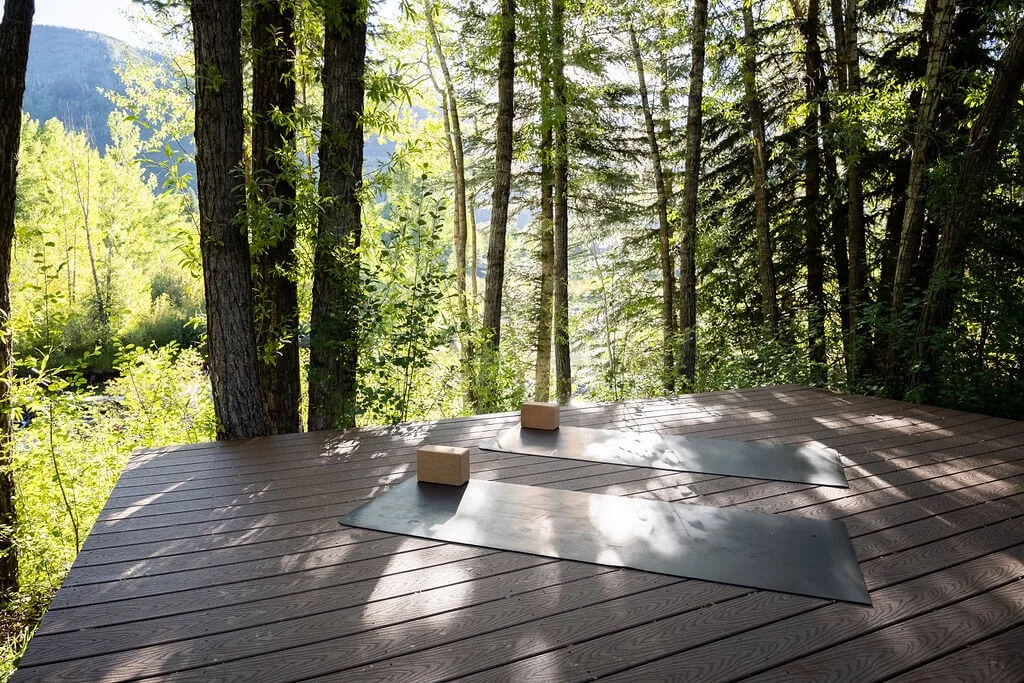 A wooden deck among trees has two exercise mats laid out in the sunlight.