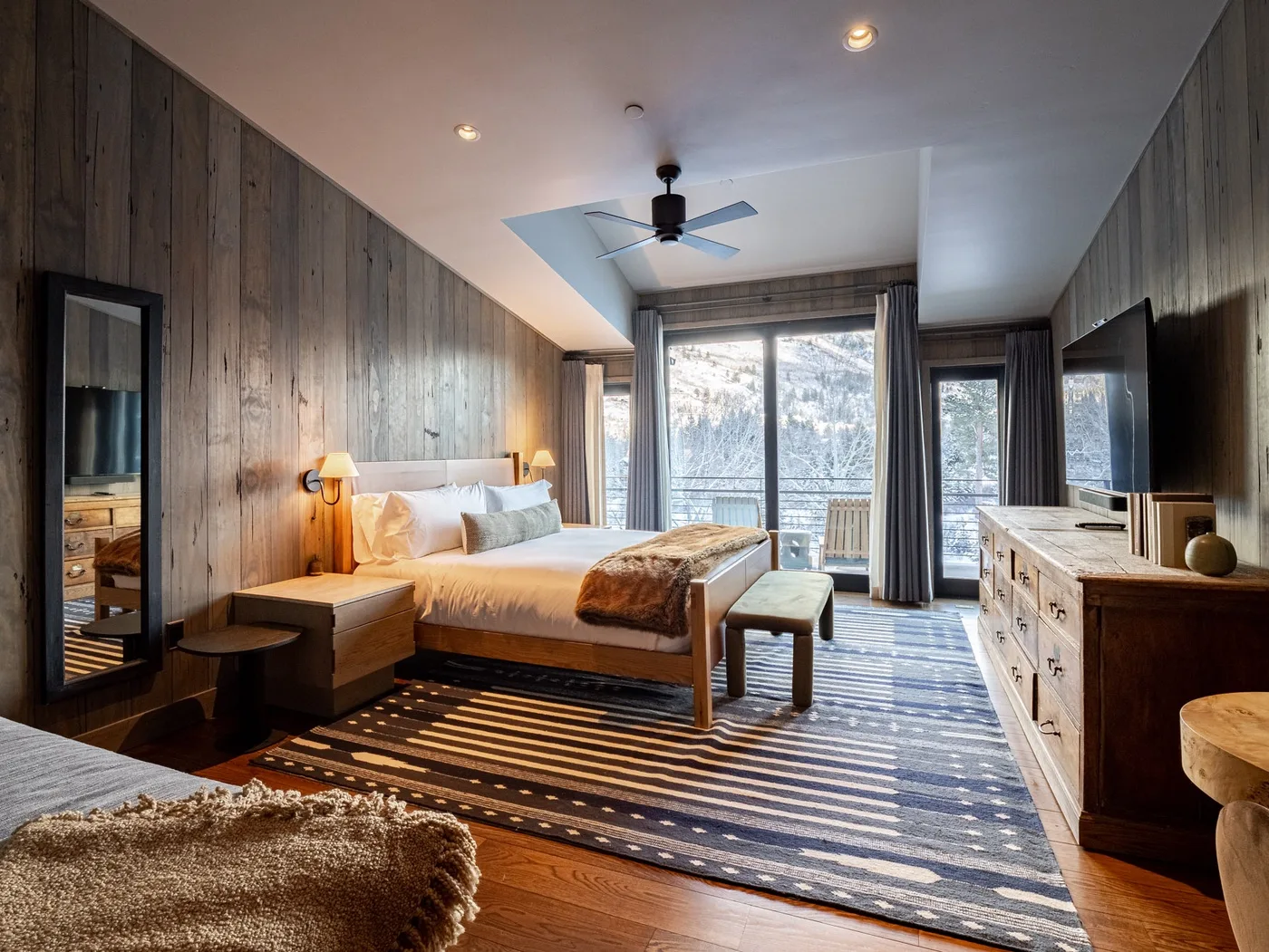 A bedroom with a bed, striped rug, and large windows.