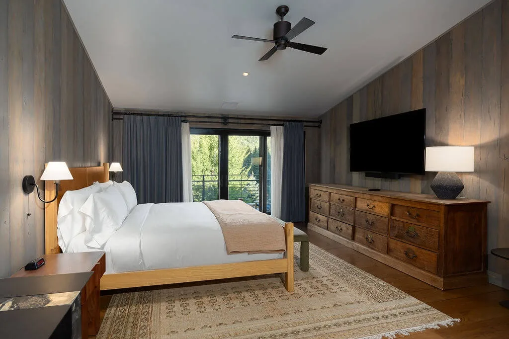 A bedroom with a bed, dresser, and large windows opening onto greenery.