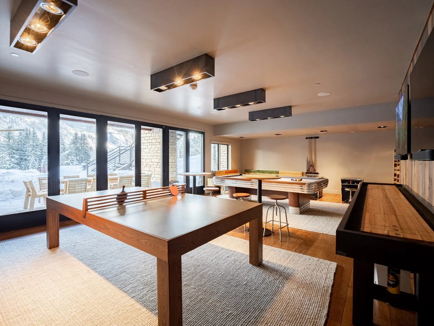 A recreation room with a large table, game tables, and windows overlooking a snowy landscape.