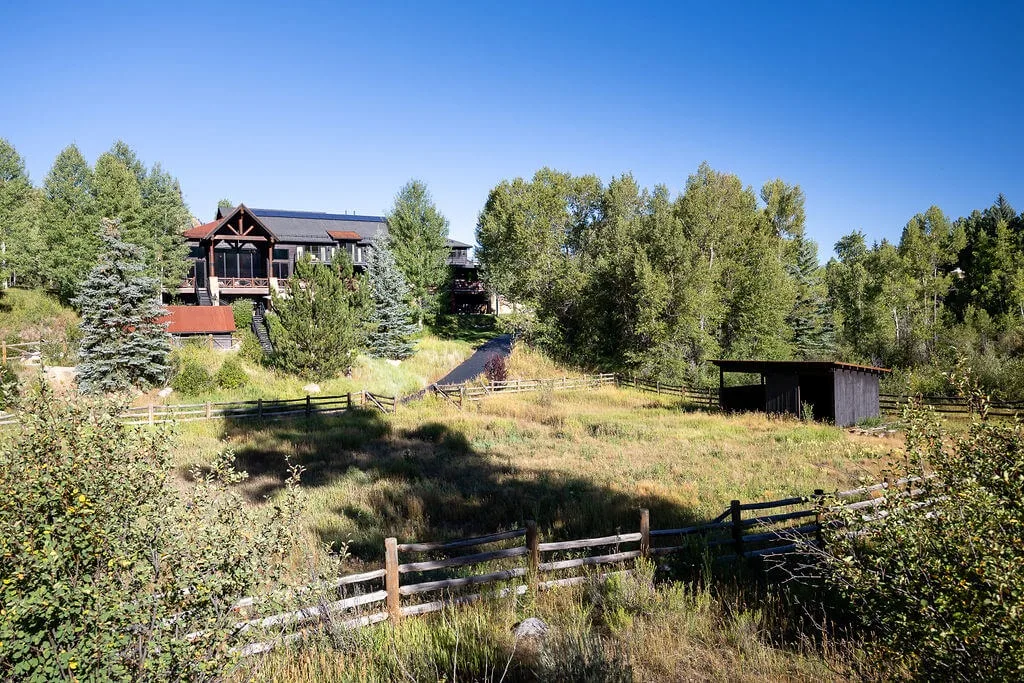 A lodge stands among trees beside an open grassy field and wooden fences.