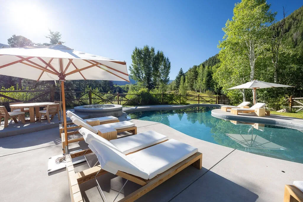 An outdoor poolside patio has lounge seating, umbrellas, and surrounding trees.