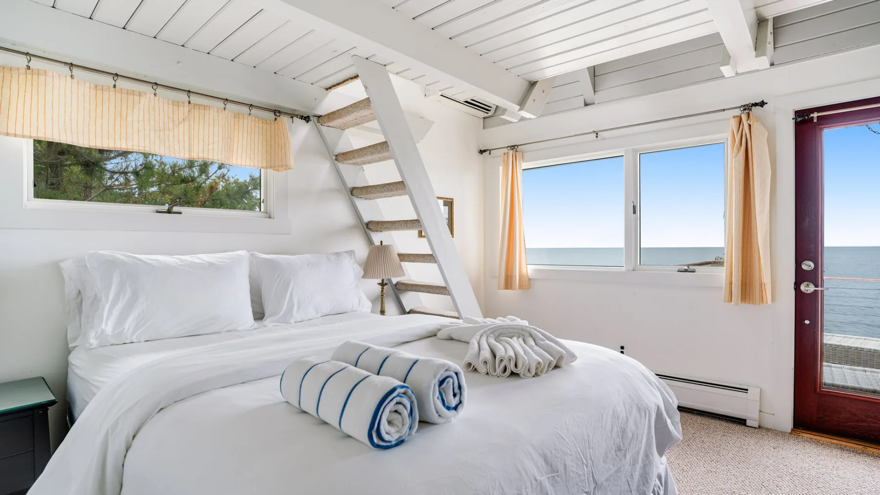 Un dormitorio luminoso con ropa de cama blanca y grandes ventanas frente al agua.