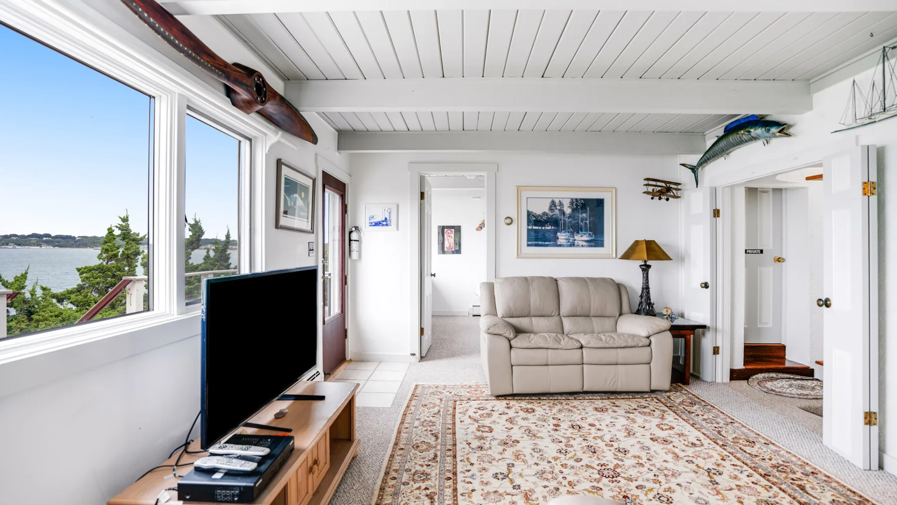 Una sala luminosa con sofá, televisor y grandes ventanas con vistas al agua.