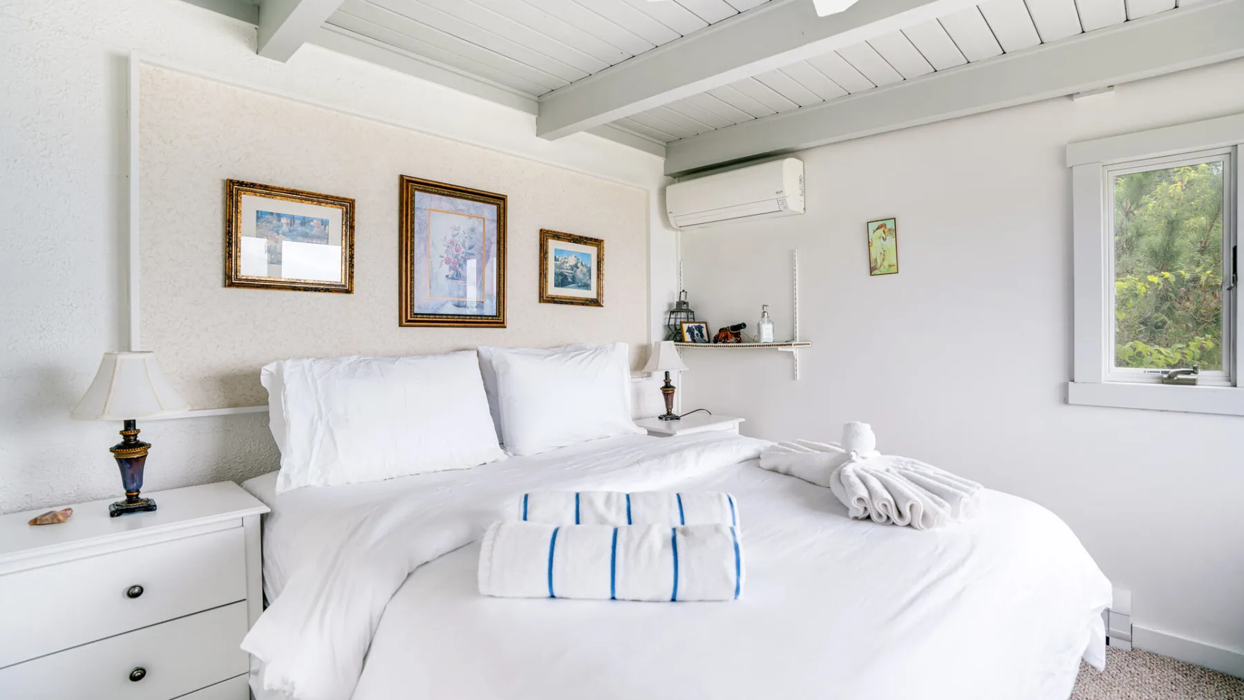 Un dormitorio luminoso con ropa de cama blanca, cuadros enmarcados y una ventana con vistas a la vegetación.