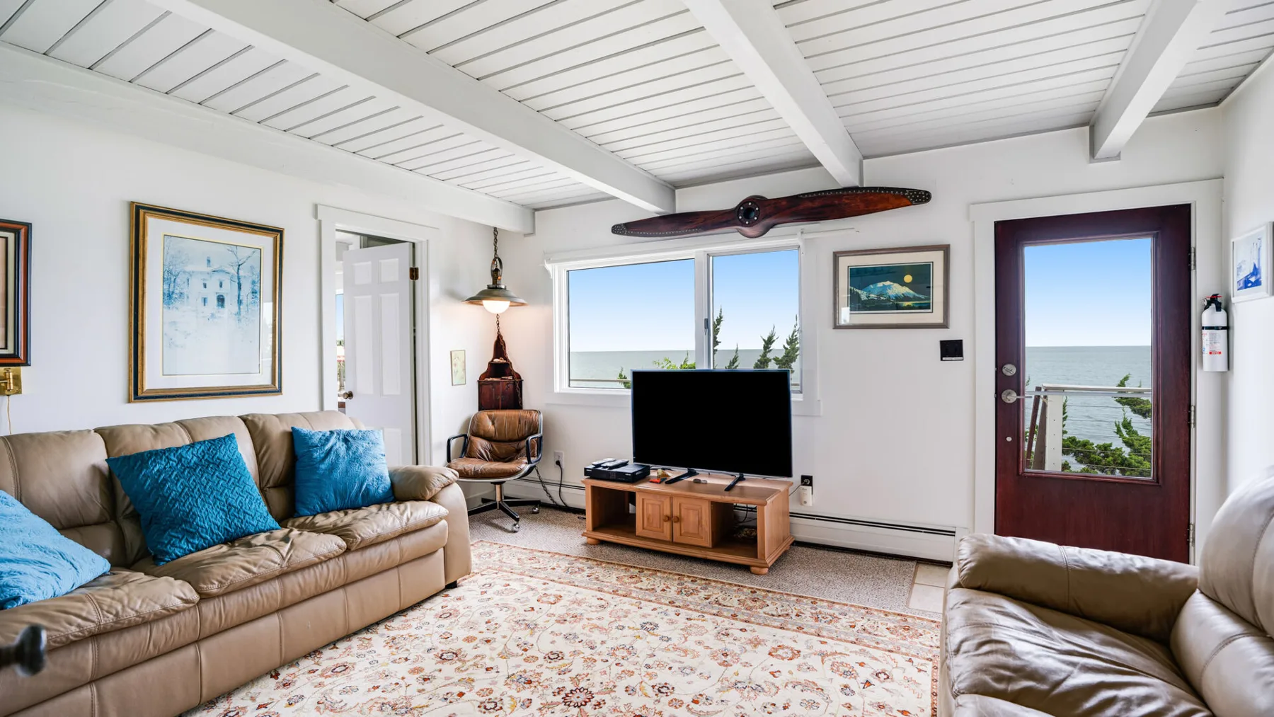 Una sala luminosa con asientos, un televisor y vistas al mar por las ventanas.