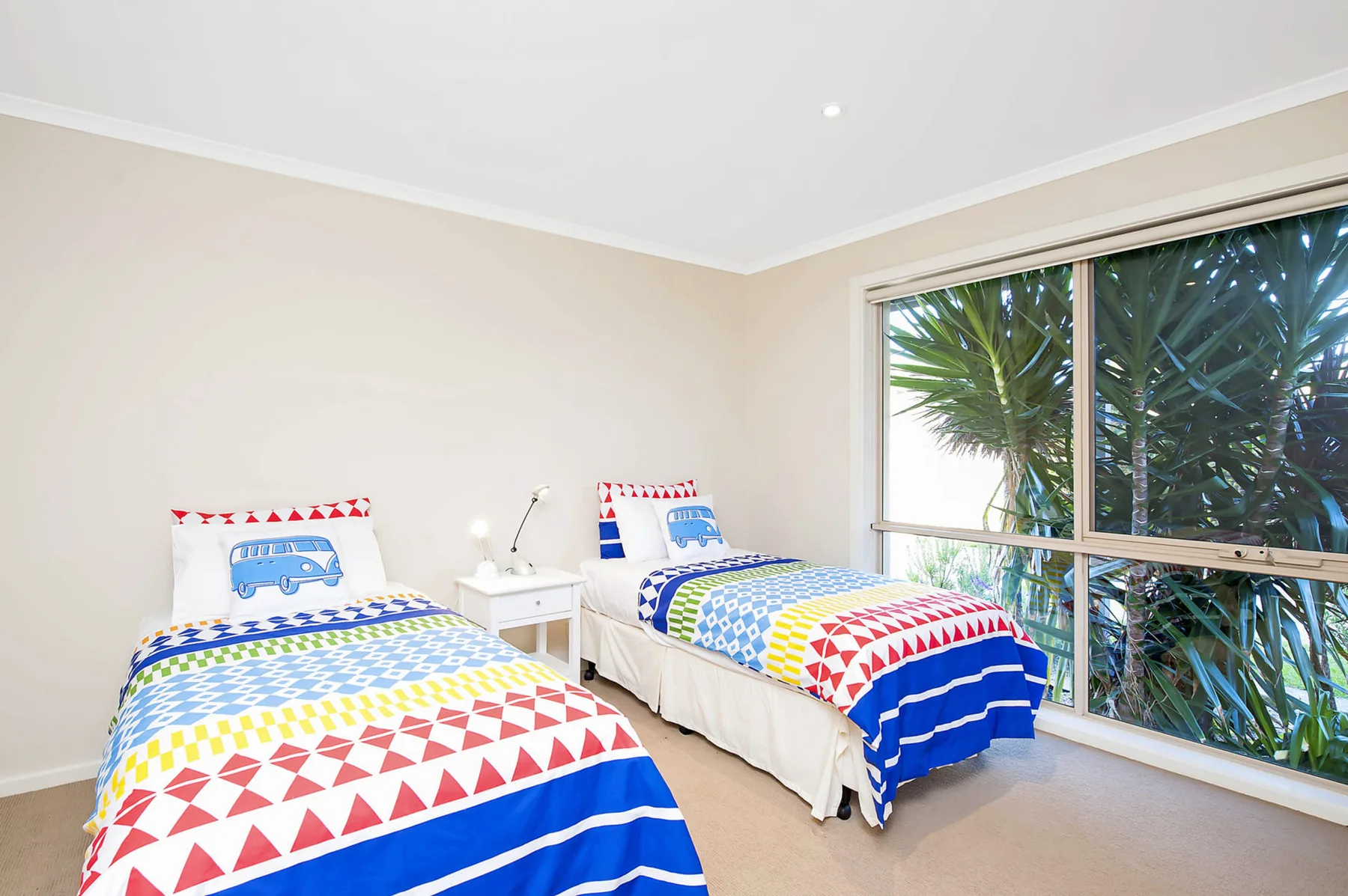 A bright bedroom with two beds beside a large window overlooking greenery.