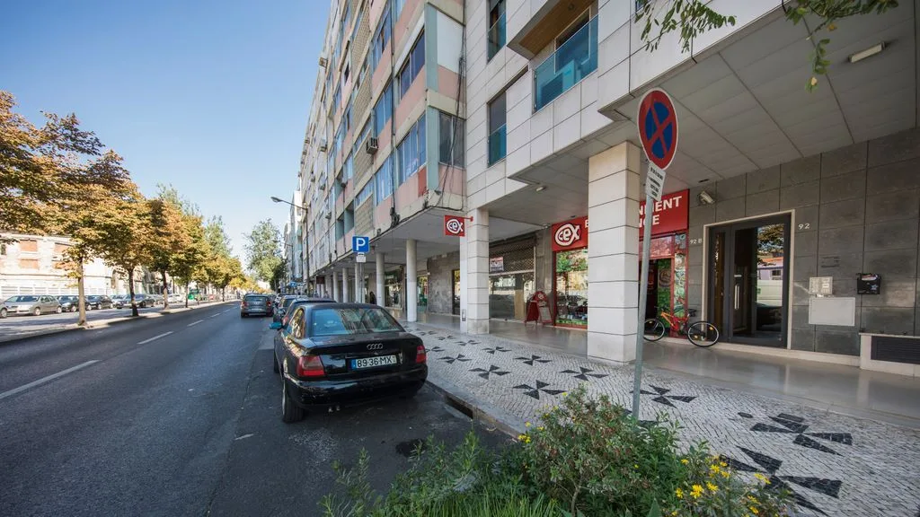 A city street with parked cars beside a modern building and storefronts.