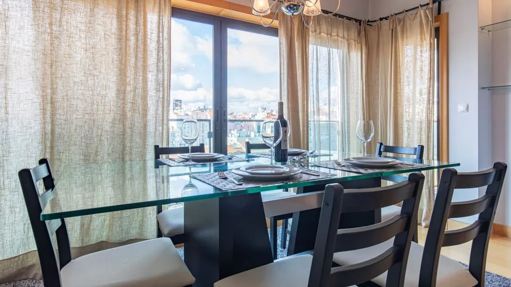 A dining area with a glass table beside large curtained windows.