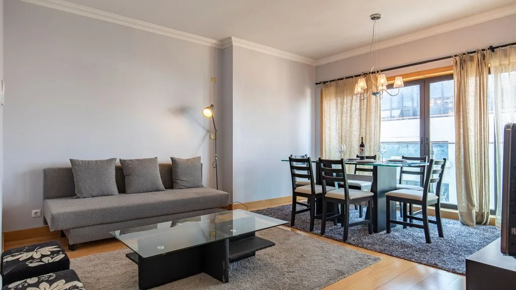 Una sala y comedor con asientos grises, una mesa de centro de vidrio y una mesa de comedor junto a las ventanas.