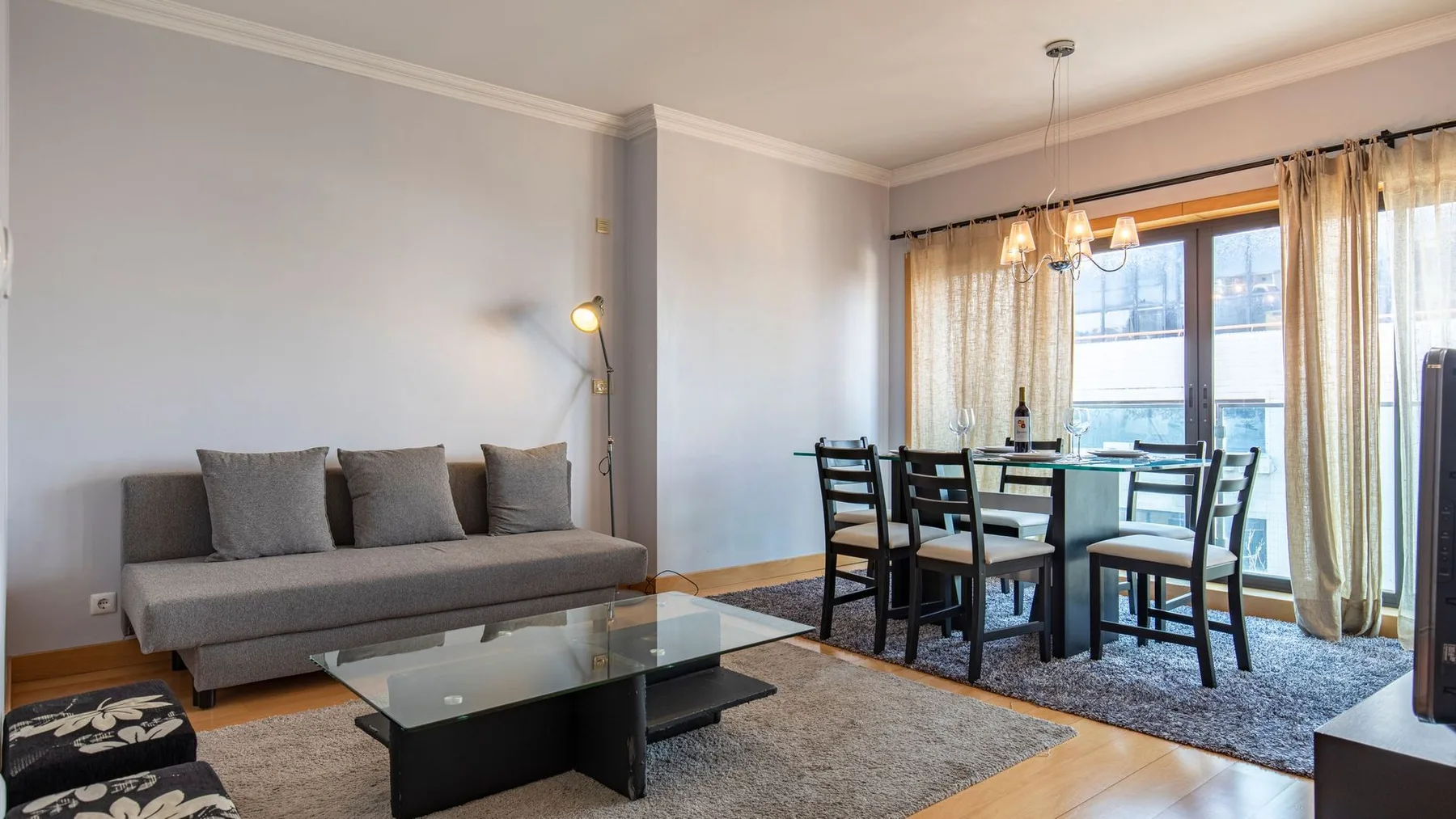 A living and dining area with seating, a glass coffee table, and large curtained windows.