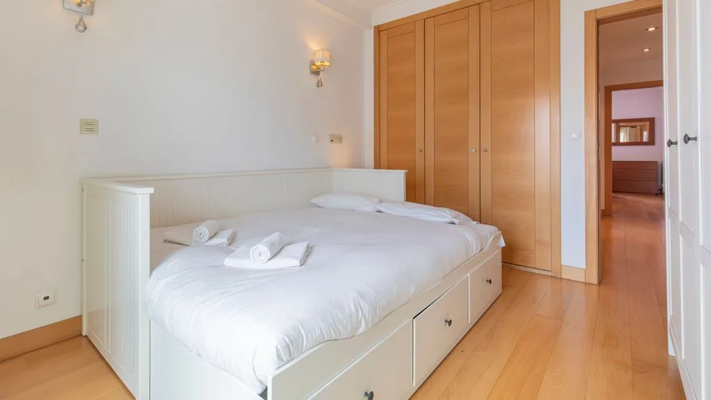 A bright bedroom with a white bed, wooden wardrobes, and an open doorway.