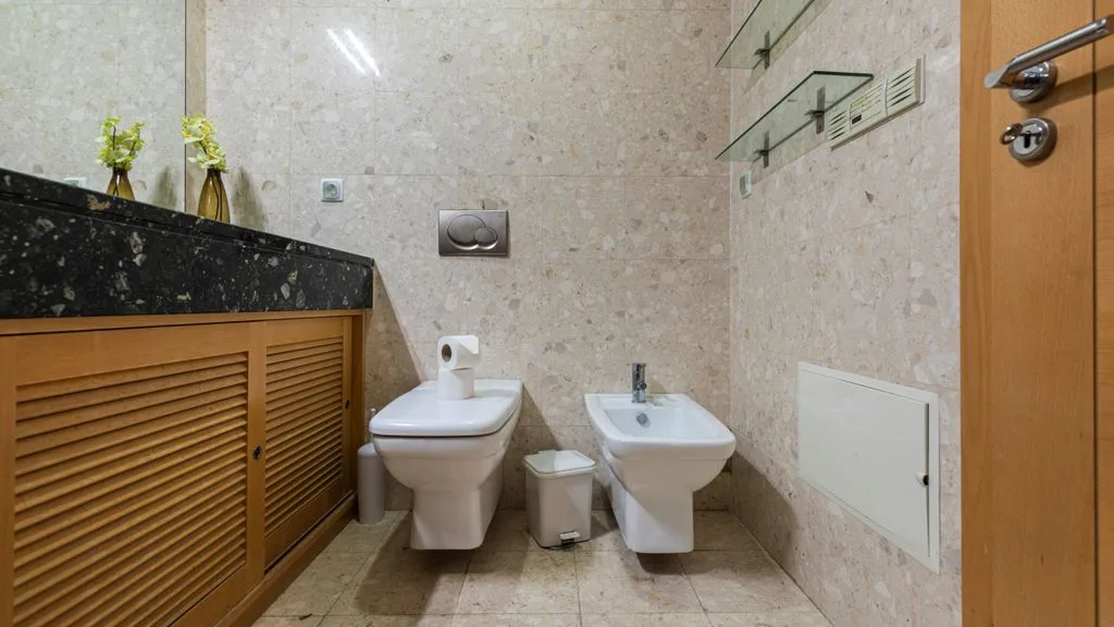 A tiled bathroom with a toilet, bidet, and wooden vanity.
