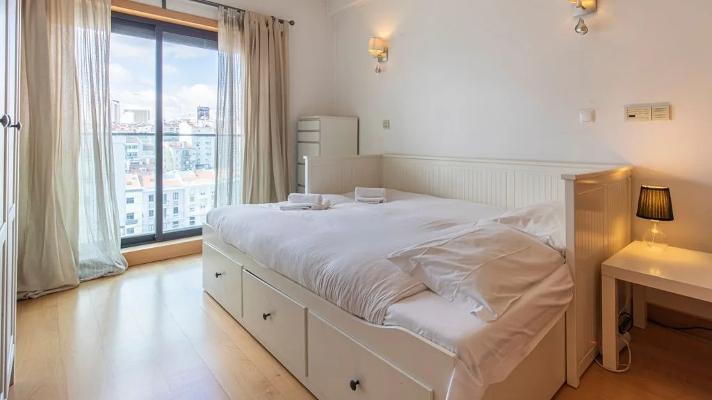 A bedroom with a large bed beside a tall window overlooking nearby buildings.