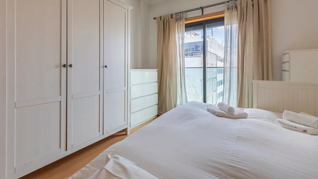 A bedroom with a white bed, wardrobes, and a window overlooking nearby buildings.