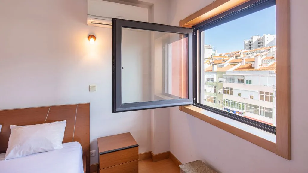 A bedroom with a bed beside an open window overlooking nearby buildings.
