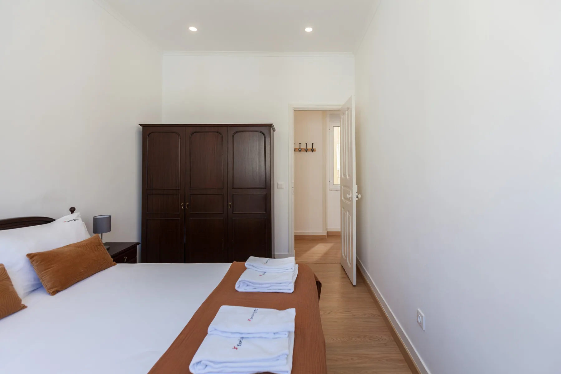 Dormitorio con una cama, toallas dobladas y un armario oscuro junto a una puerta abierta.