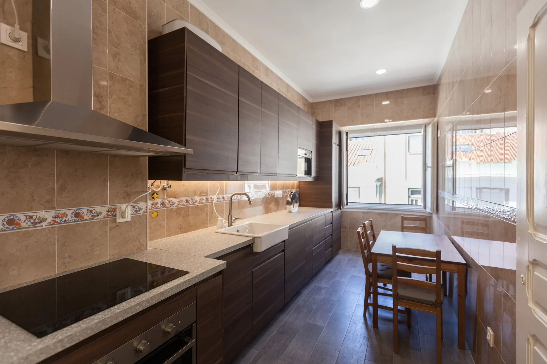 Una cocina alargada con armarios oscuros, una placa de cocción y una mesa junto a las ventanas.