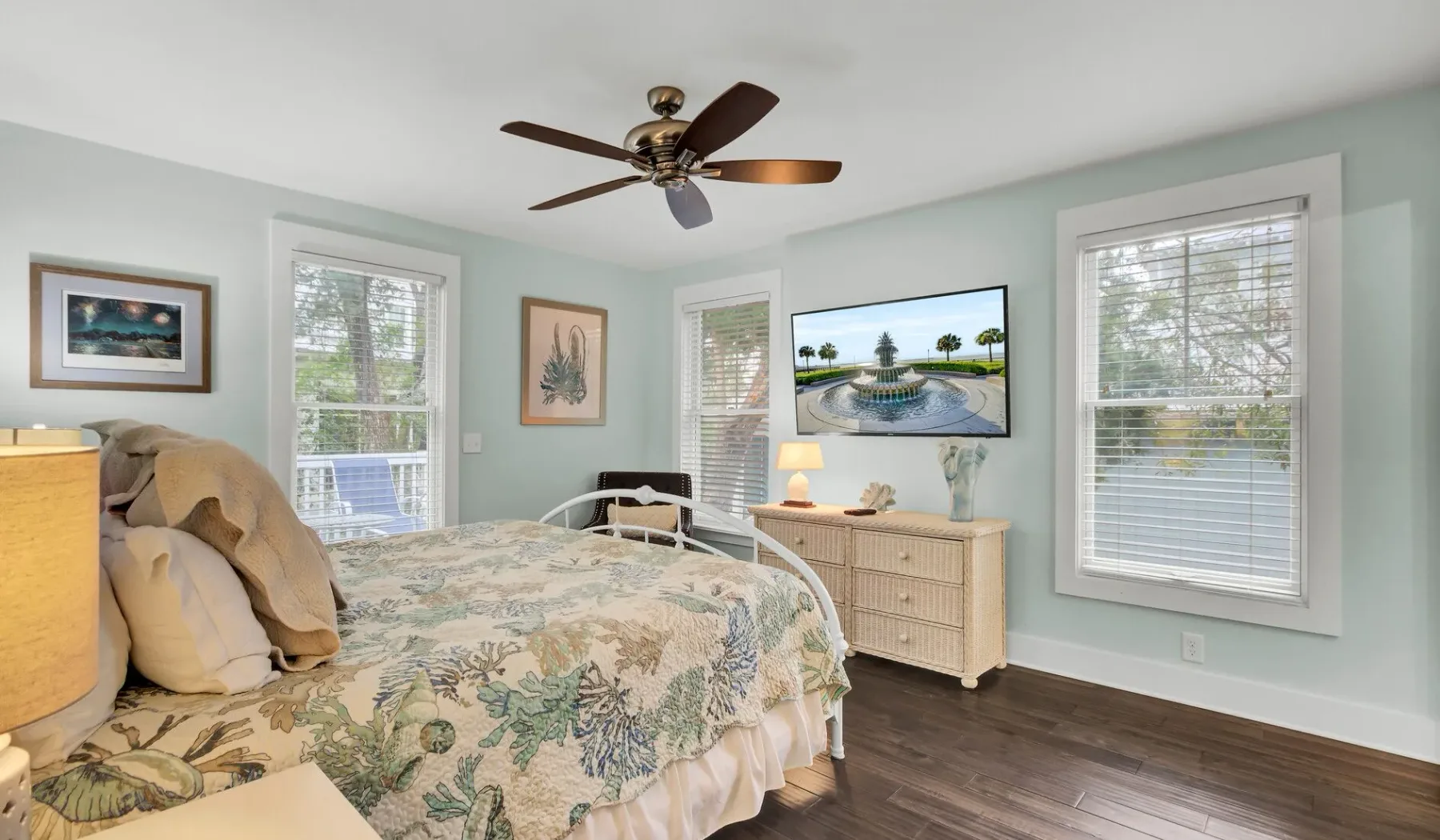 A bedroom with a floral bedspread, pale walls, and windows overlooking greenery.