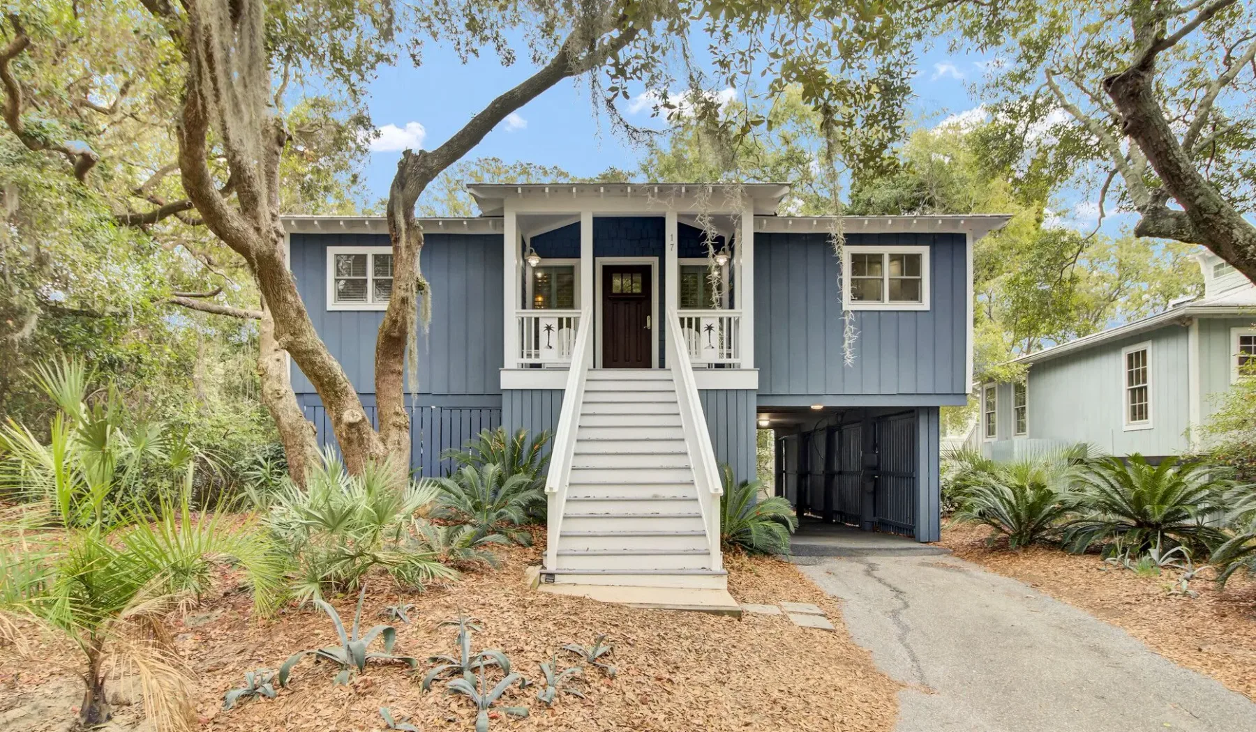 A raised blue house with a white staircase stands among leafy trees.