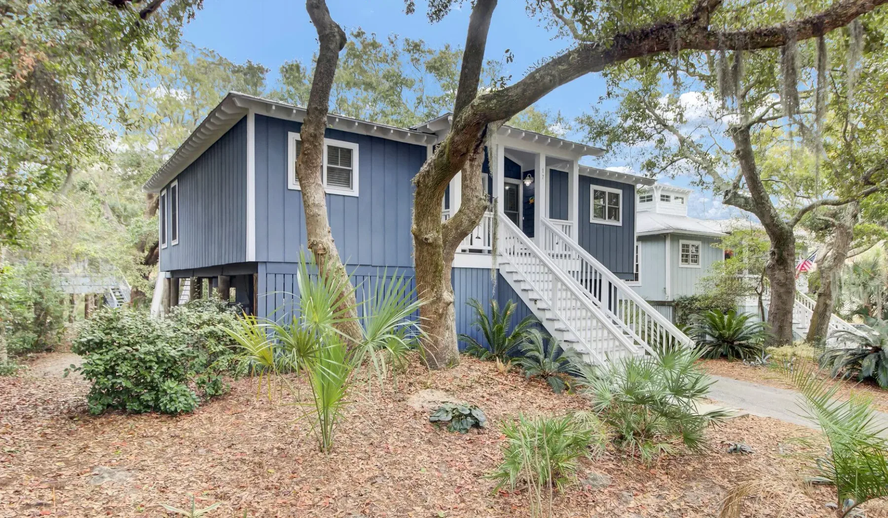 A blue raised house with white stairs stands among trees and low landscaping.