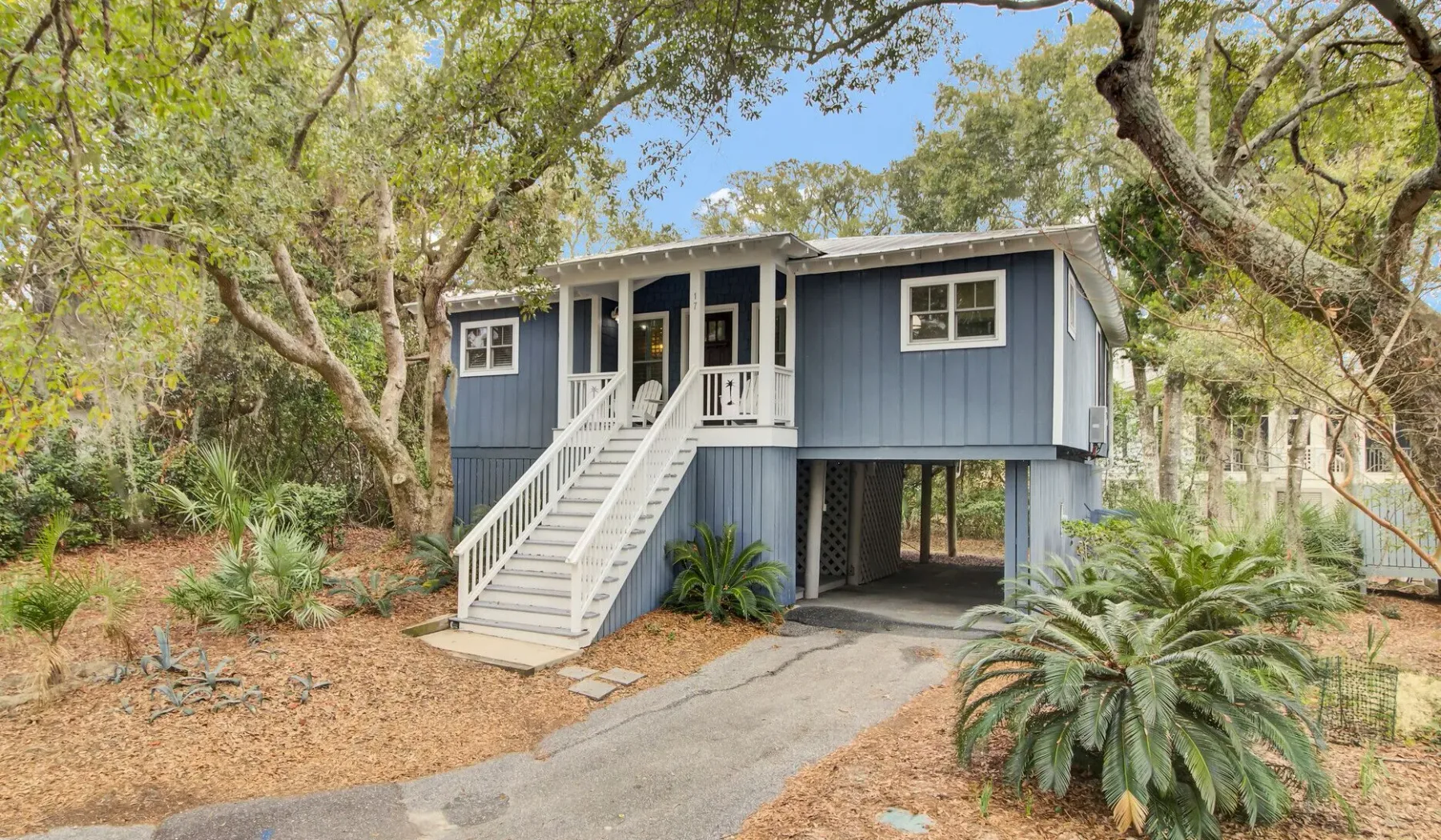 A raised blue house with white stairs stands among trees beside a paved driveway.