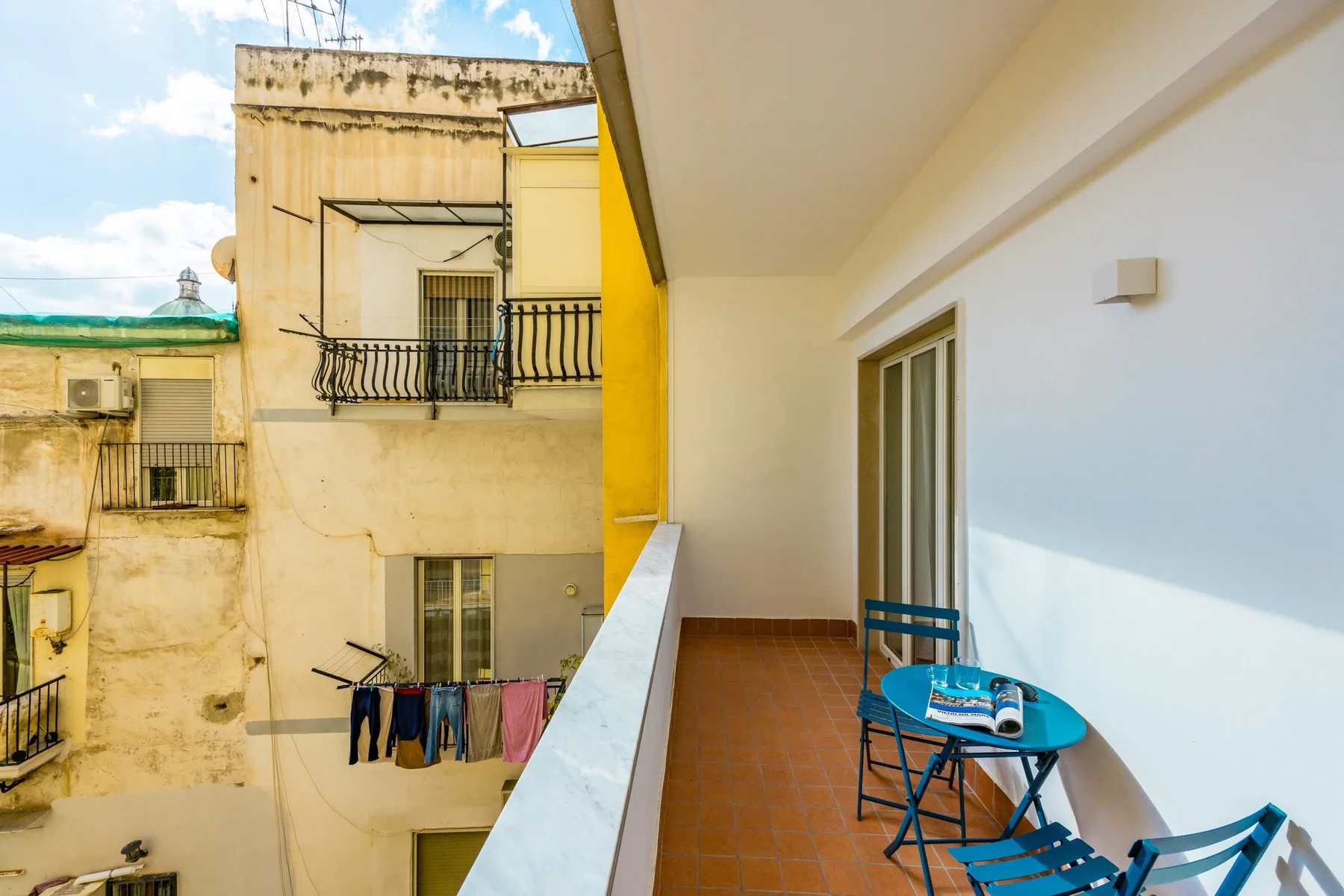 A narrow outdoor balcony with blue seating and a small table overlooks neighboring buildings.