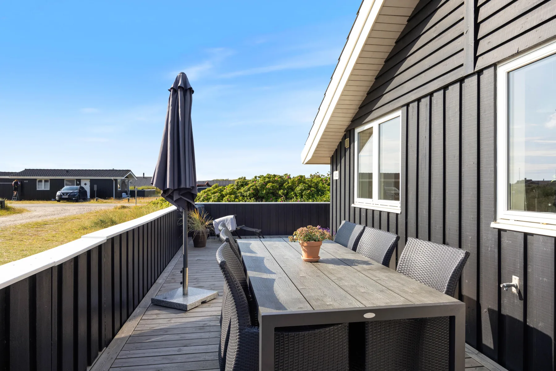 Una terraza exterior tiene una mesa de comedor, asientos y una sombrilla cerrada junto a una casa.