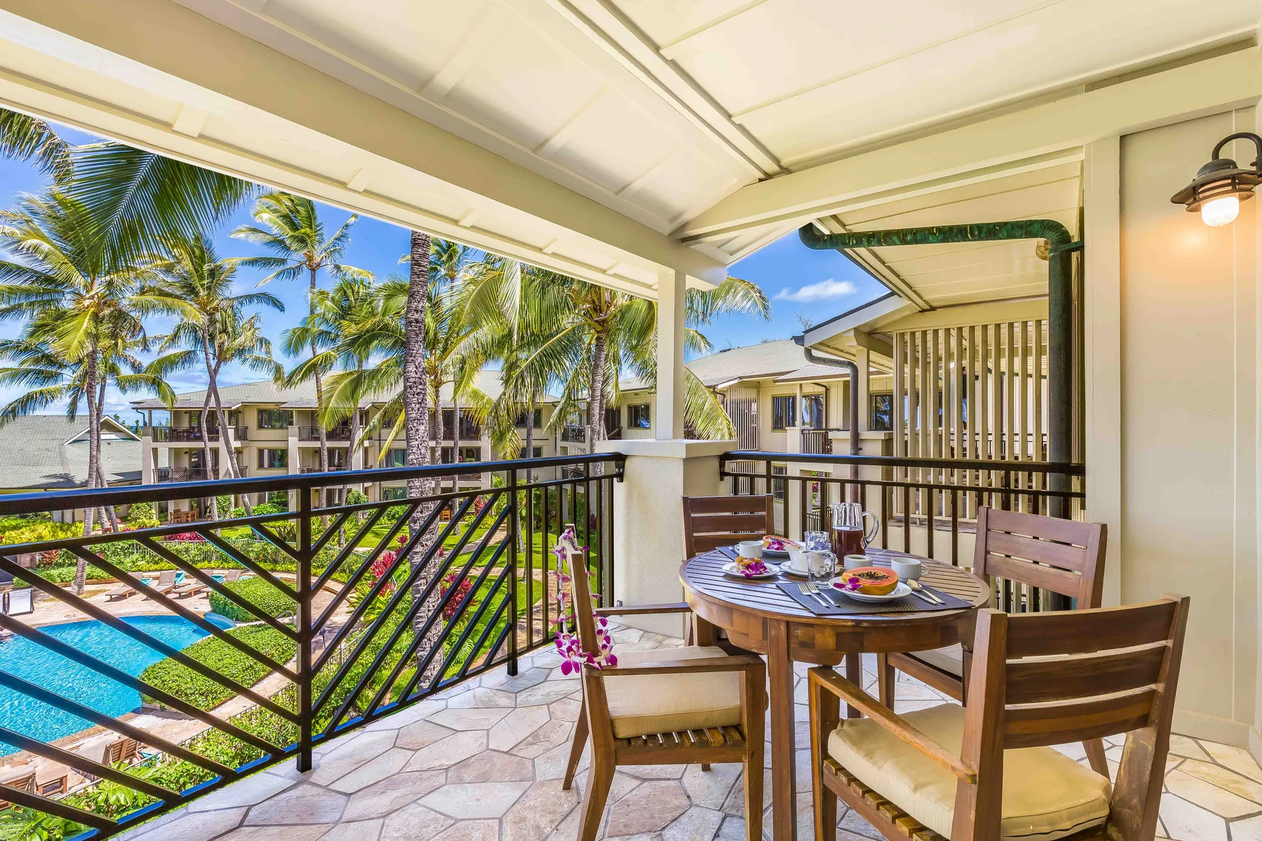 A covered balcony dining area with a table and chairs overlooks palm trees and a pool.