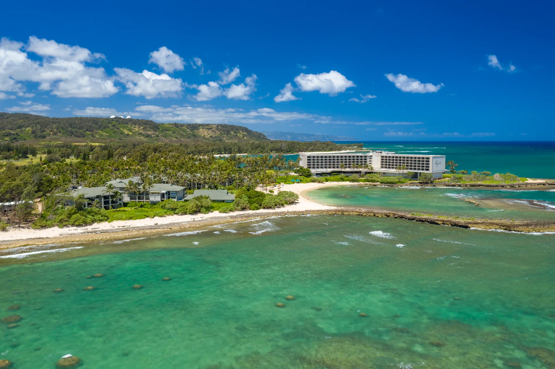 A coastal resort complex sits beside a sandy beach and clear turquoise water.