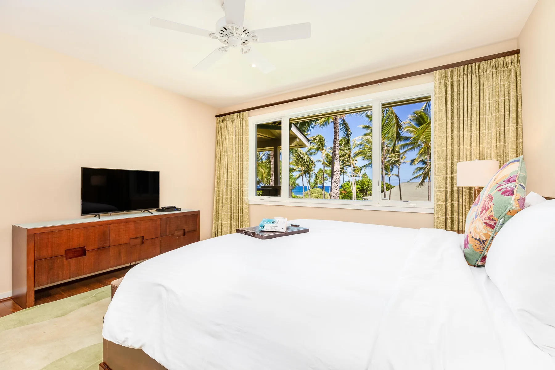 A bedroom with a large bed, a television, and palm trees visible through the window.