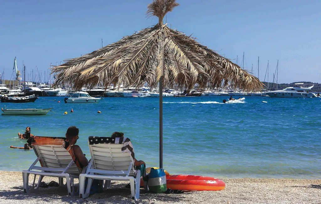 Una escena de playa con asientos bajo una sombrilla de paja, frente a embarcaciones en el agua.