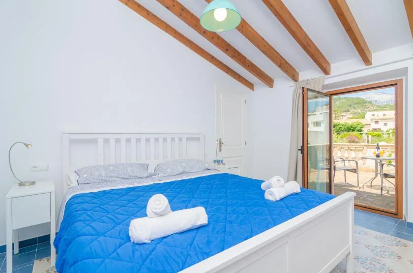 A bright bedroom with a bed covered in a blue quilt beside an open balcony door.