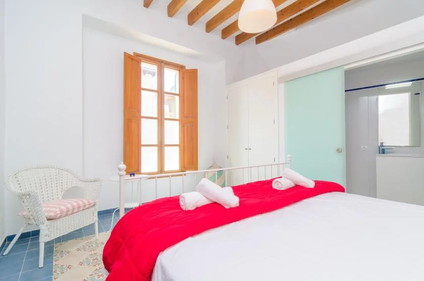 A bright bedroom with a red bedspread, a wooden-framed window, and exposed ceiling beams.
