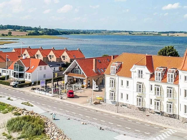 A row of waterfront buildings with red roofs faces a road and a broad body of water.
