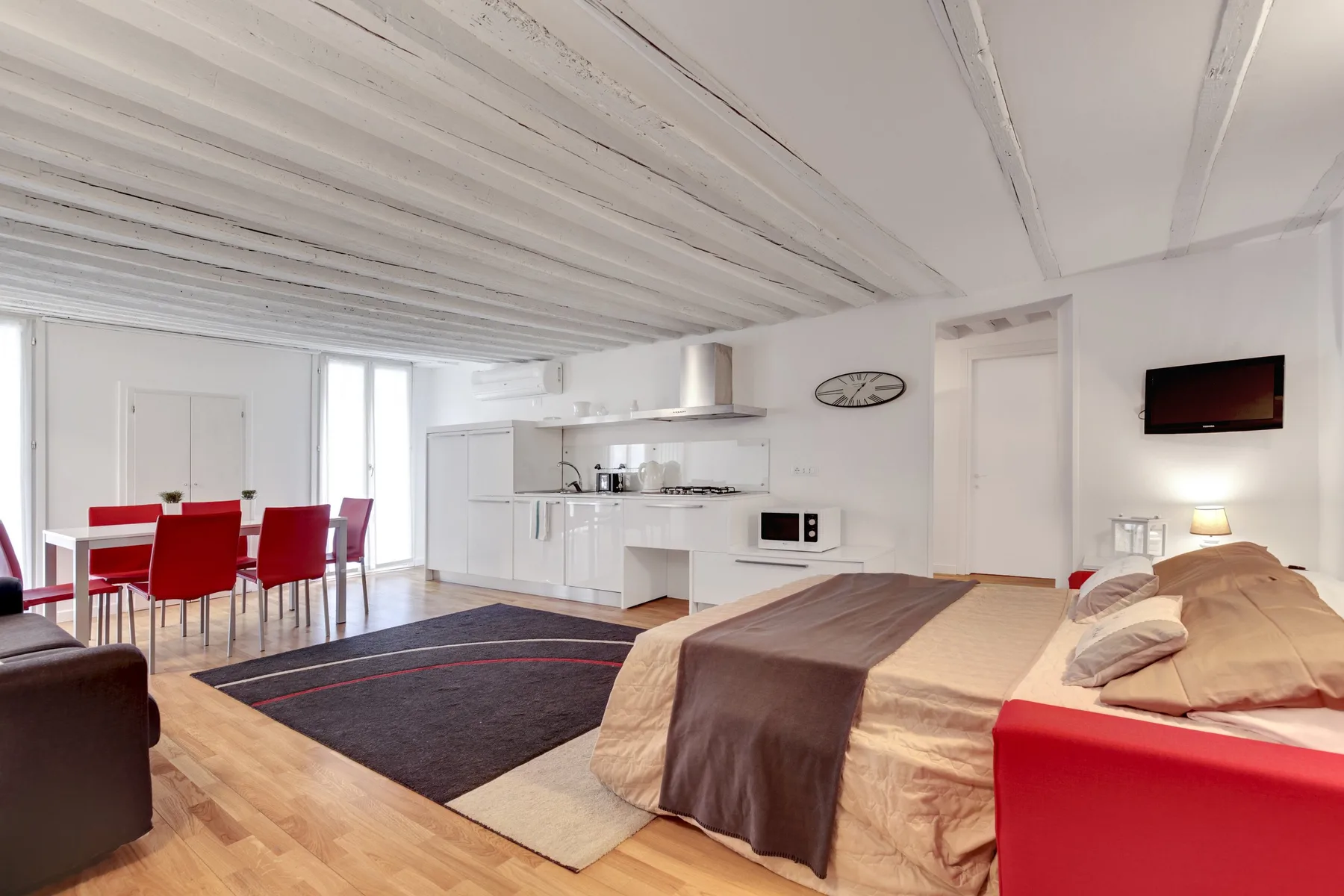 Una zona abierta de dormitorio y cocina incluye una cama, una mesa de comedor y una alfombra oscura.