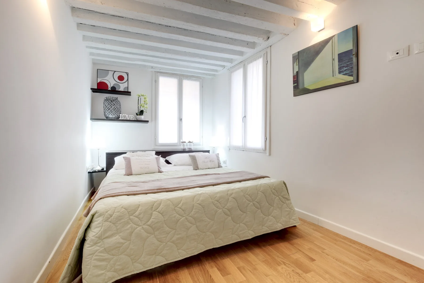 Un dormitorio luminoso con una cama bajo vigas blancas y ventanas.