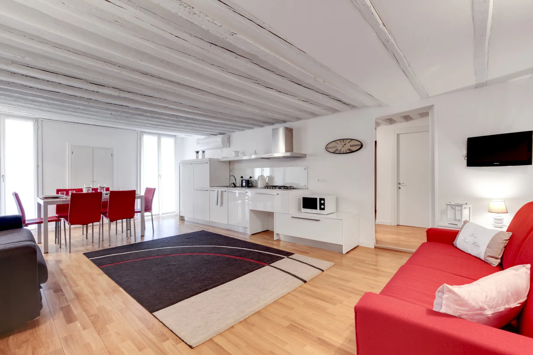 Una sala de estar diáfana con sofá rojo, mesa de comedor y cocina blanca.