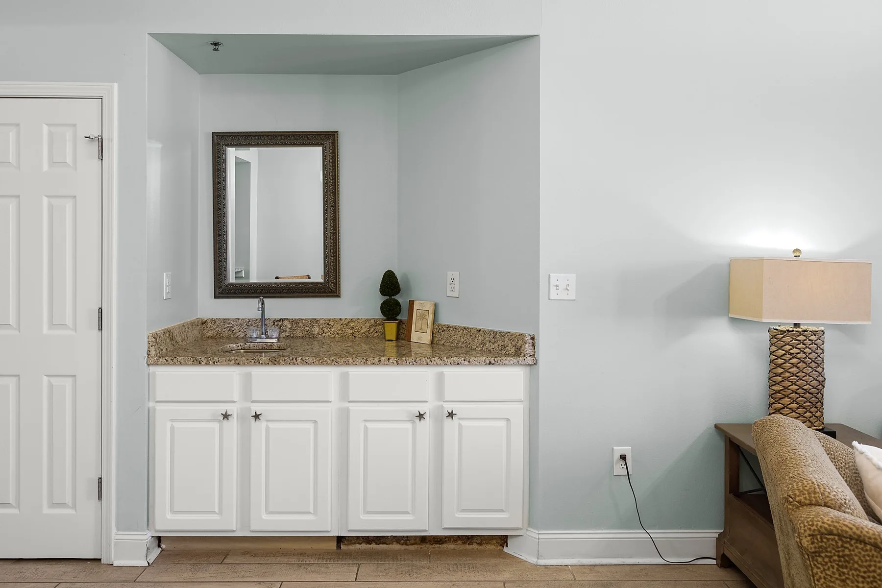 A bathroom vanity with a mirror and sink stands beside a bed and lamp.