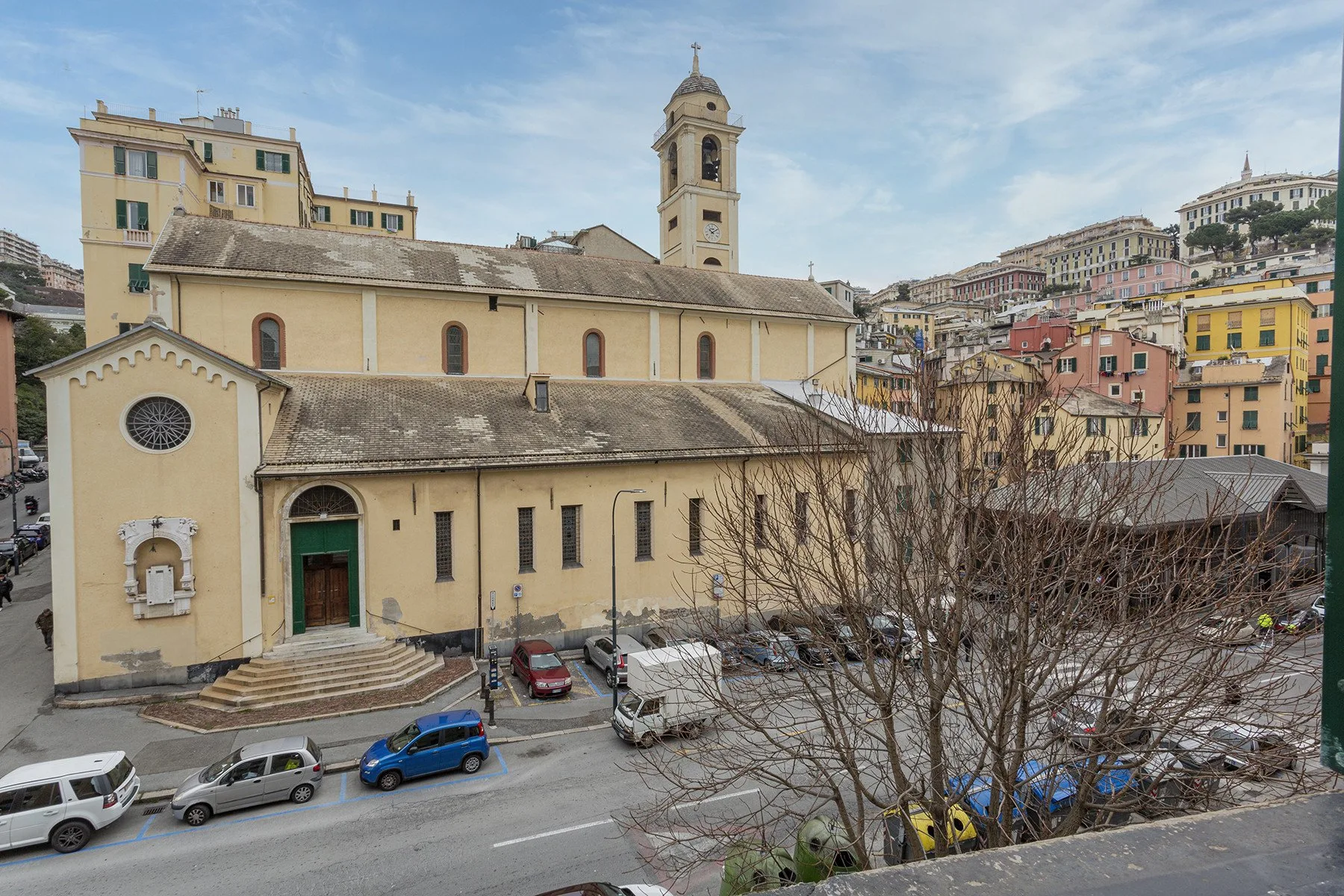 Una iglesia de tonos claros con campanario se alza junto a una calle con autos estacionados.