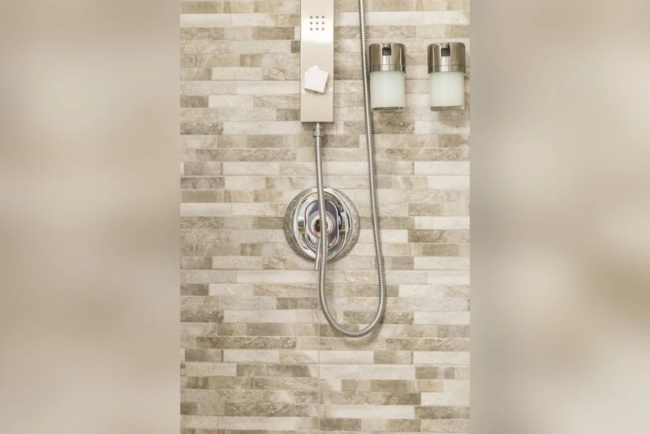 A tiled shower wall with a handheld showerhead and two dispensers.
